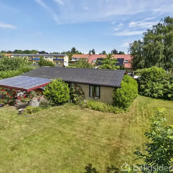 Et lille hus med en sorttag er omgivet af en velplejet græsplæne og tæt bevoksning. En veranda med rød blomsterdekoration grænser op til huset i en stille forstadskontekst.