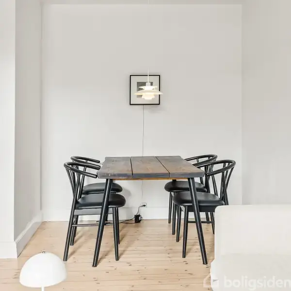 Et træbord omgivet af fire sorte stole. En hvid lampe hænger over bordet, mens en lille ramme dekorerer væggen i det minimalistisk indrettede rum.