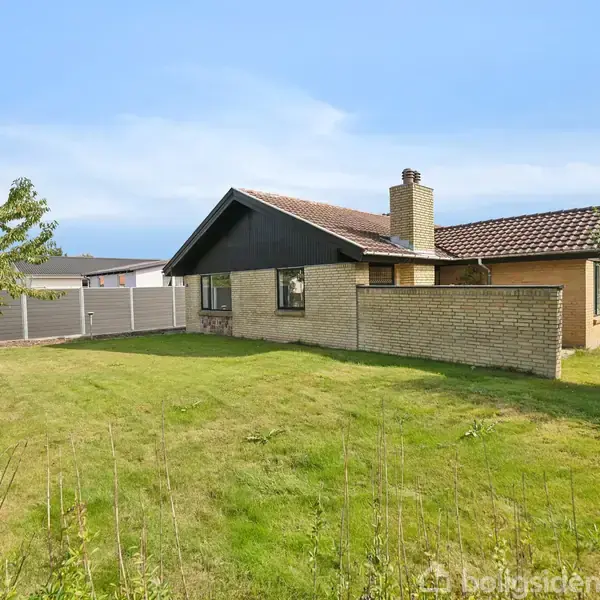 Et enfamiliehus med gul murstensfacade står stille på en velplejet græsplæne. I baggrunden ses en garage og et hegn, og himlen er blå.