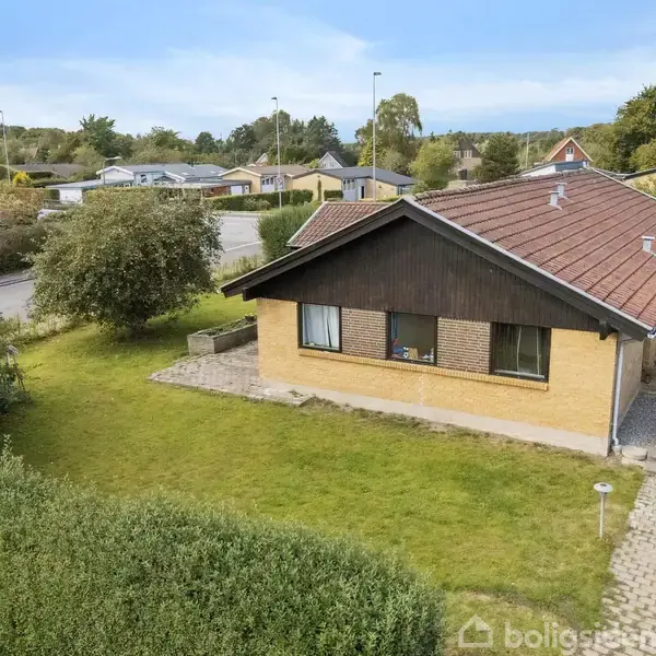 Et gult murstenshus med rødt tag og store vinduer står stille på en velplejet græsplæne omgivet af en hæk, i et boligkvarter med flere huse i baggrunden.