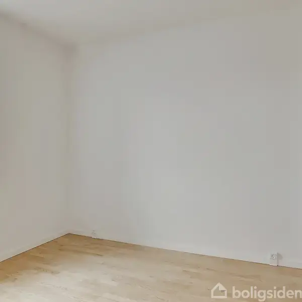 Et tomt rum har lyse trægulve. En enkelt stor vindue lader lys strømme ind, mens hvide vægge omslutter rummet og en radiator er placeret under vinduet. Ingen tekst til stede.