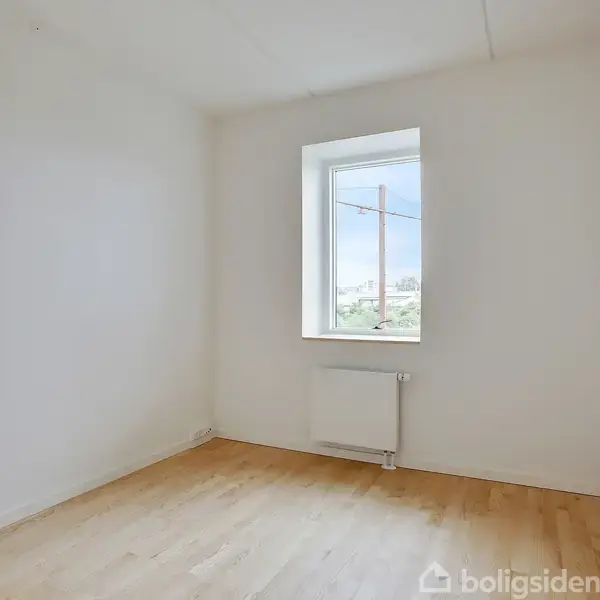 Et tomt rum med lyse trægulve har et vindue, der viser en udsigt til træer og himmel. Der er en radiator under vinduet, og væggene er hvide.