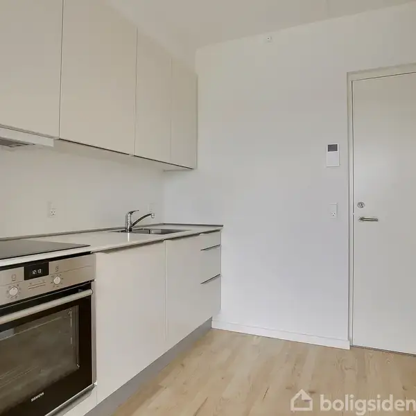 Køkken med hvide skabe og en indbygget ovn til venstre. Et hvidt bord med vask løber langs væggen. Trægulv og en lukket hvid dør til højre.