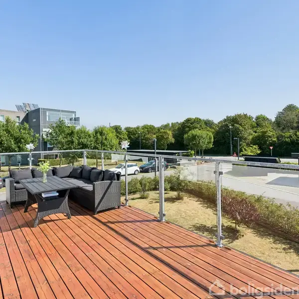 Terrasse med træplanker indeholder sorte havemøbler. Terrassen har glasrækværk og udsigt over en vej og grønne træer i et byområde under en klar blå himmel.