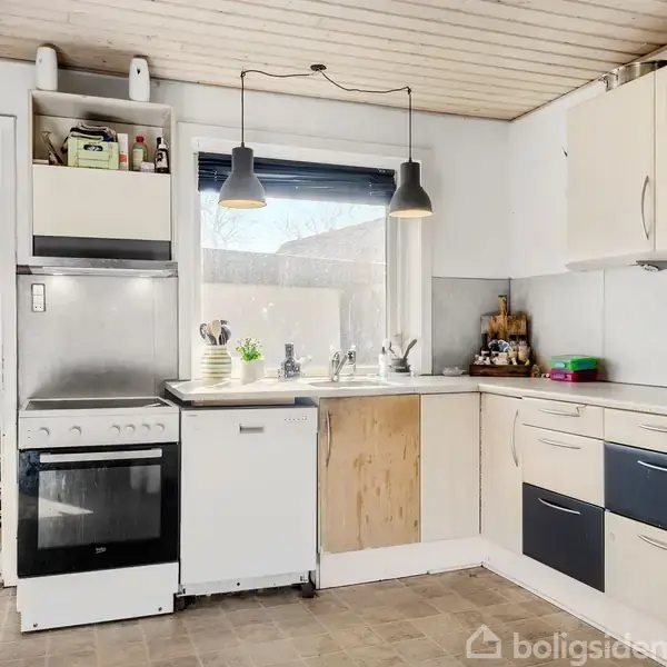 En køkkenvask med to lamper hængende over, omgivet af hvide skabe og en ovn i et lyst køkken. I baggrunden ses en entré med jakker og sko.