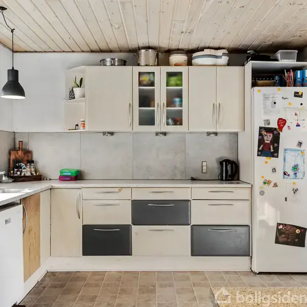 Et køkken med hvide og grå skabe langs væggen. Køleskabet er dekoreret med magneter og billeder. Frugtskål med appelsiner står på gulvet. Lyse trælofter og flisegulv.