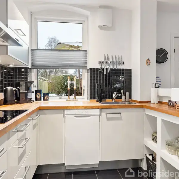 Køkken med hvide skabe og træbordplader. En sort flisevæg omgiver et vindue, og forskellige køkkenredskaber står fremme. Køkkenmaskiner og opvaskemaskine ses på bordet.