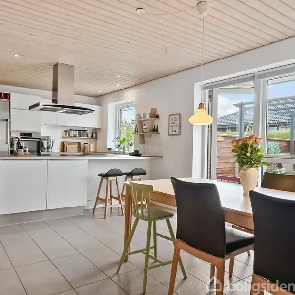 Køkken-alrum med spisebord og stole i forgrunden, køkkenø med barstole i midten. Rummet er lyst med store vinduer og hængende lamper. Terrassedør leder ud til en udendørs terrasse.