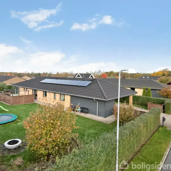 Et rækkehus med solpaneler står på en velplejet have med en trampolin, legeredskaber og en hæk. I forgrunden ses en vej med parkerede biler og et fortov.