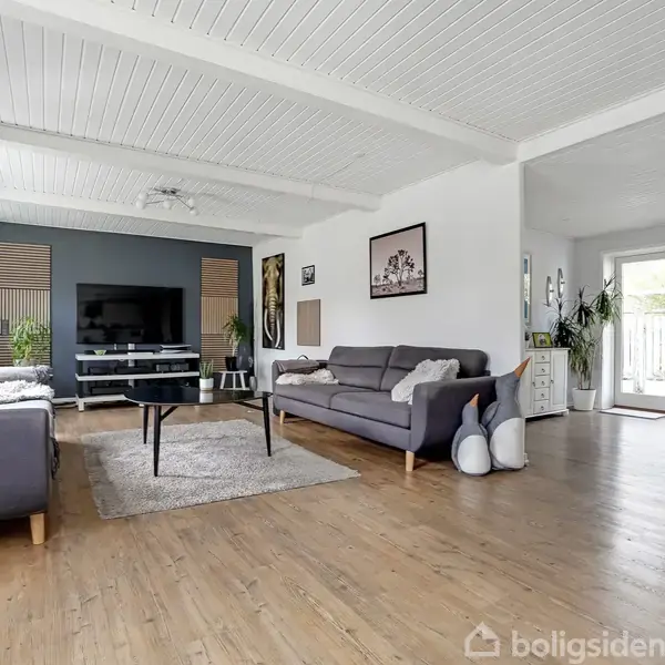 Soveværelset har en seng med blåt sengetøj, placeret mod væggen. To vinduer med planter står på den ene side, mens en garderobe og et spejl er på den anden.