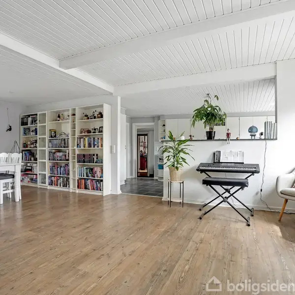 Et moderne køkken med hvide skabe og sorte fliser. Et spisebord med grå stole står i midten. Lysindfald fra vinduer og forskellige køkkenredskaber og planter ses på bordpladen.