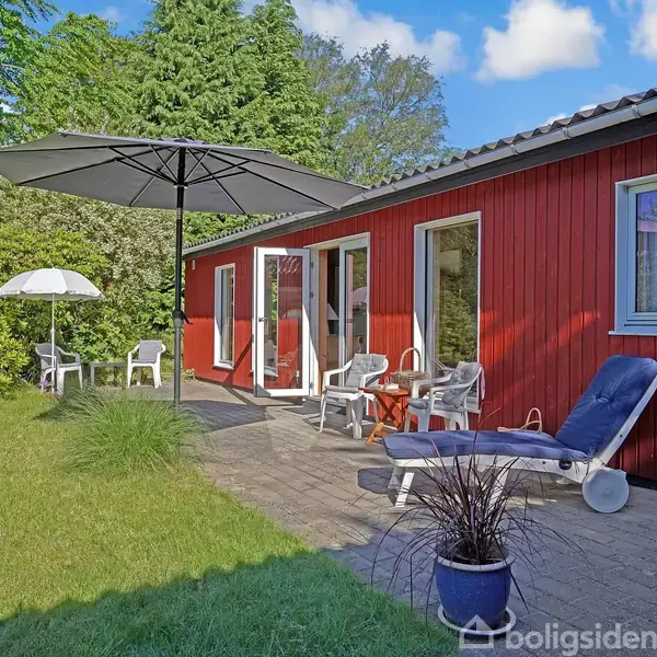 Et rødt træhus med store vinduespartier åbner mod en terrasse, hvor havemøbler og parasoller står. Terrassen ligger i en grøn have omgivet af træer og buske.