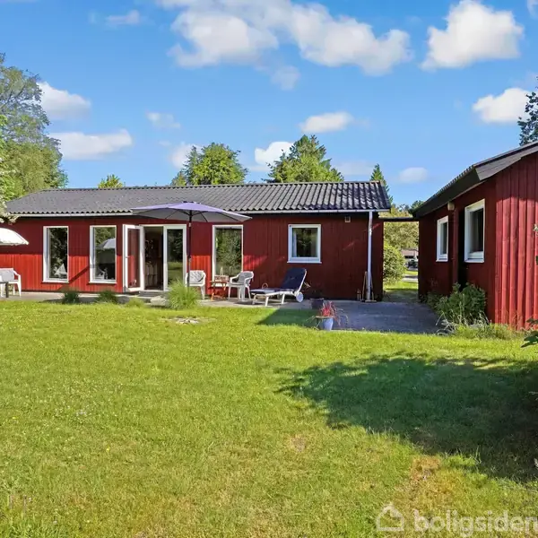 Rødt træhus står i en grøn have med græsplæne, omgivet af træer og buske. Havemøbler med parasoller er placeret på terrassen foran huset, under en blå himmel med spredte skyer.