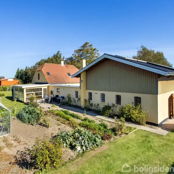 Gulmalet hus med rødt tag står blandt frodig have med drivhus. Omkranset af grønne træer under en klar blå himmel.