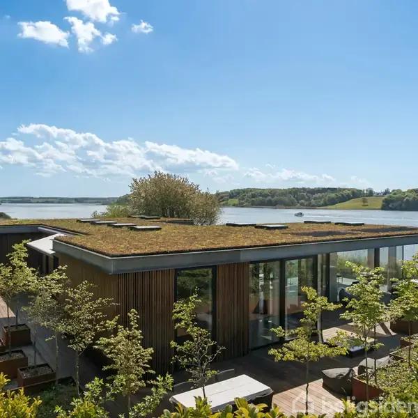 Moderne hus med grønt tag står stille ved en sø, omgivet af frodig natur og træer, under en klar blå himmel med spredte skyer.