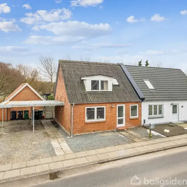 En murstensvilla udgør hovedfokus, med en grå tilbygning. Villaen står stille ved en vej med en tilknyttet carport til venstre, omgivet af en åben have og vintertræer.