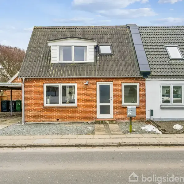 Rødt murstenshus med gråt halvtag og hvide vinduesrammer. Foran huset er der en indkørsel med grus og en carport. Husnummer og postkasse er synlige i den lille forhave.