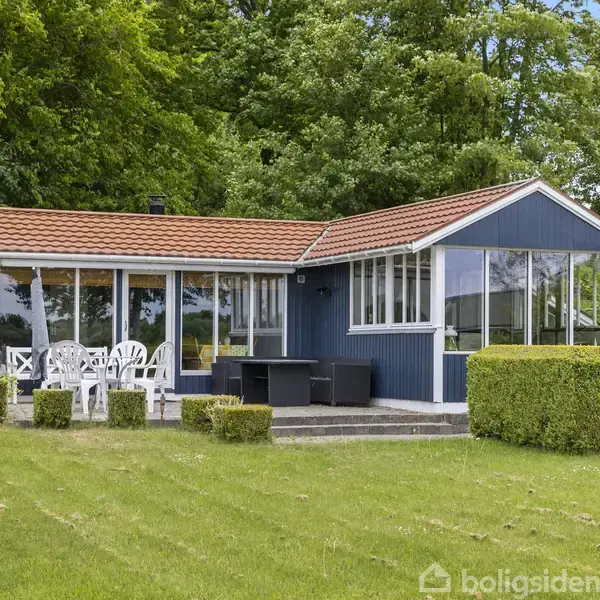Et blåmalet sommerhus med rødt tegltag står i en grøn have. Der er en terrasse med hvide og sorte havemøbler omgivet af hæk og store træer i baggrunden.