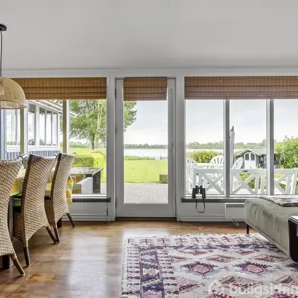 Spisebord med flettede stole står i stue med store vinduer, der viser udsigt til have og vand. En sofa til højre, en hængende lampe, og gulvtæppe under.