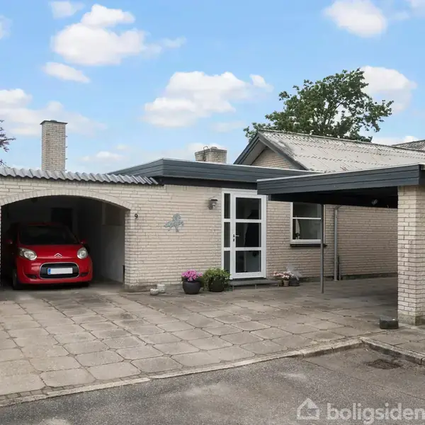 En bil holder parkeret i en carport ved siden af et murstenshus med fladt tag. Flere potteplanter står på flisebelagt terrasse. Skyfrit, med træer i baggrunden.