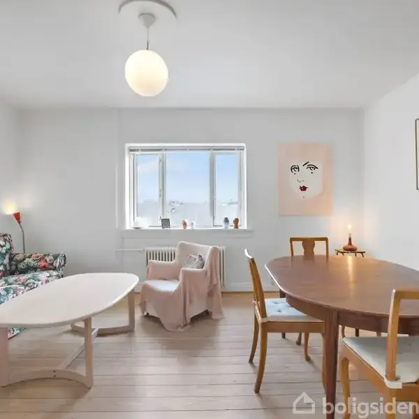 Et lyst, minimalistisk stueområde med en blomsterfarvet sofa og rundt træbord. Et hvidt ovalt sofabord og en enkelt lyserød lænestol. Vægkunst inkluderer en enkel ansigtstegning.