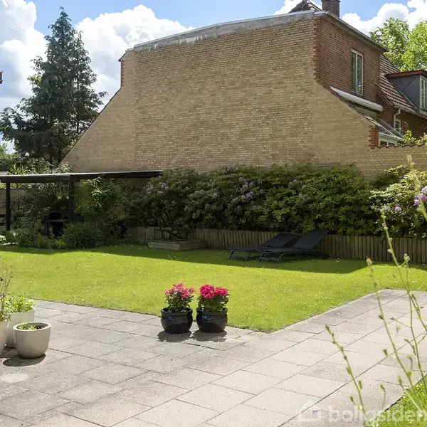 Terrasse med fliser og blomsterkrukker; solsenge står på græsplæne ved murstenshus. Grønne buske og træer omkranser området. Skyerne er synlige på den blå himmel.