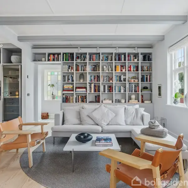 Sofa dominerer rummet foran en reol fyldt med bøger. To træstole med læderpuder står på hver side af et lavt bord. Store vinduer lader naturligt lys ind.