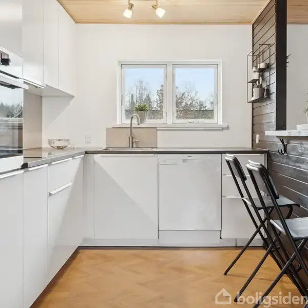 Et moderne køkken med hvide skabe og apparater. To høje stole står ved en mørk trækant. Vindue viser træer udenfor. Lys brænder over en hvid opvaskemaskine.