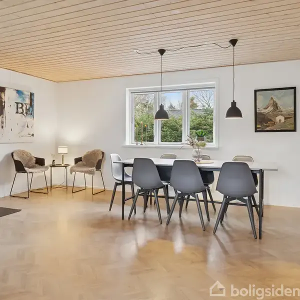 Pejsen er tændt og står i et lyst, moderne stuerum med trægulv og træloft. Til venstre ses to stole og et lille bord, mens et spisebord med stole er foran et vindue.