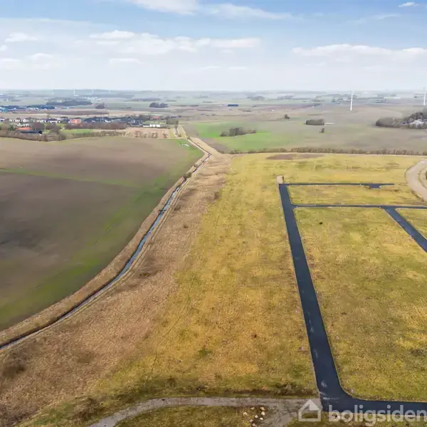 En ubebygget mark er opdelt i græsarealer med asfalterede veje. Landskab i baggrunden viser spredte træer og vindmøller under en blå himmel.