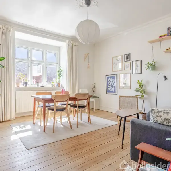 En stue med et spisebord og stole i midten, omgivet af store vinduer. Rummet har planter, hylder med bøger og en grå sofa. Væggene er dekoreret med kunst og billeder.
