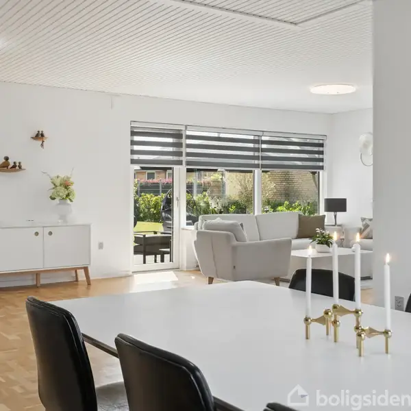 Spisestue med hvidt bord og sorte stole, stearinlys og vægur på væggen. Sofa og skænk står ved vinduer med udsigt til have.