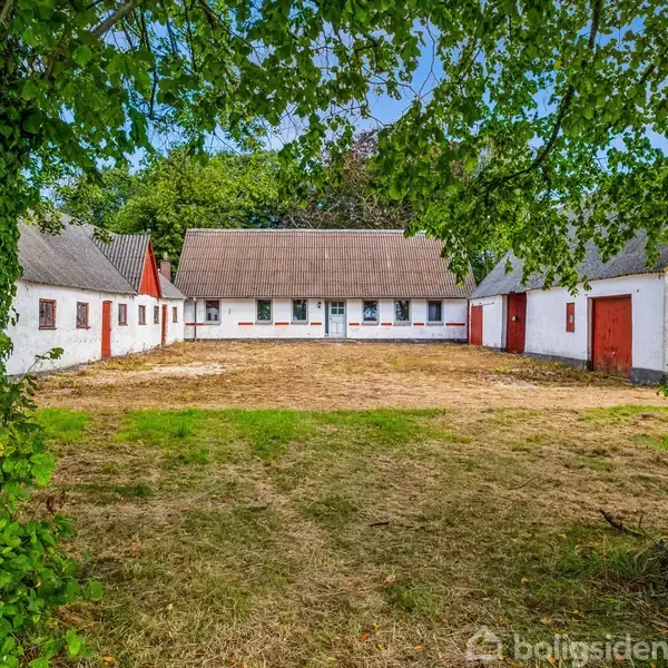 En hvid gård med røde døre og vinduer er omgivet af træer. Den ligger på en åben, græsklædt gårdsplads med en overhængende grenramme, der danner en naturlig bue.