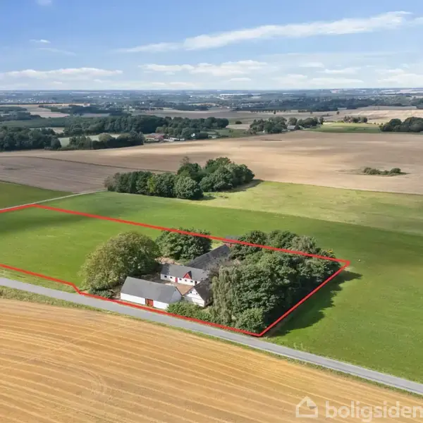 Hus omgivet af træer, ligger inden for en markeret, indhegnet område på en stor, landlig mark med spredte træer og åbne vidder i baggrunden under en blå himmel.