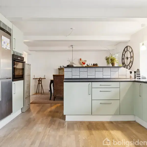 Køkken med lysegrønne skabe og sort bordplade står ud mod en spisestue. På væggen hænger et stort ur, og planter pynter skillevæggen. Lyse trægulve i hele rummet.