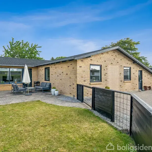 Et gult murstenshus med sort tag ligger i en velplejet have. Der er en terrasse med havemøbler og parasol til venstre, omgivet af græsplæne og indkørsel.