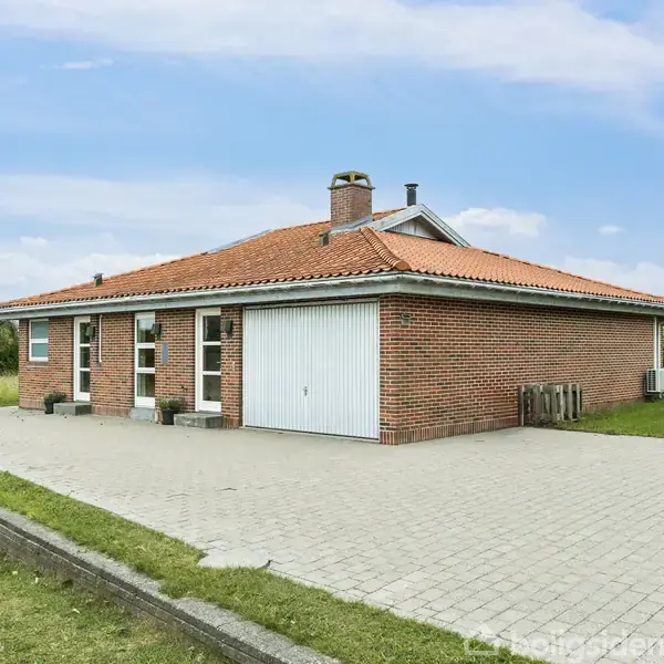Rødt murstenshus med fladt tag står stille på en stor flisebelagt terrasse. Græsplænen omgiver huset, mens træer ses i baggrunden under en klar blå himmel.