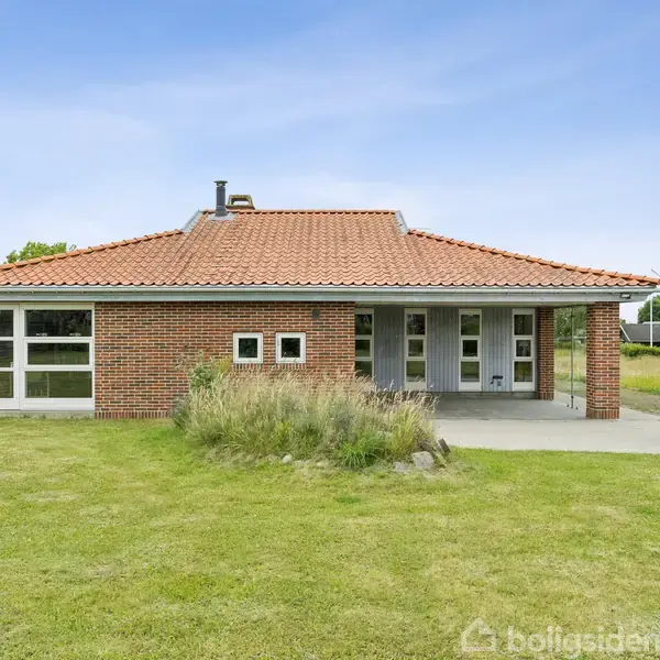 Murstenshus med rødt tegltag står stille på en grøn græsplæne. Terrassen har siddemøbler, og omgivelserne inkluderer træer og buske under en klar blå himmel.