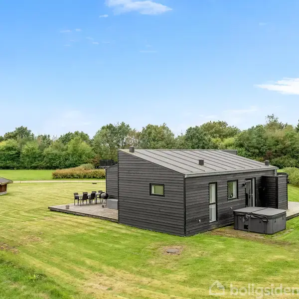 Et sort, moderne sommerhus står på en stor, velplejet græsplæne med terrasse, havemøbler og en trampolin. Omgivet af frodig natur og grønne træer under en blå himmel.