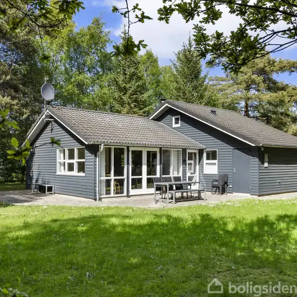 Et gråt sommerhus står midt i en grøn have omringet af træer. På terrassen foran huset står havemøbler, og sollys filtrerer gennem løvet.