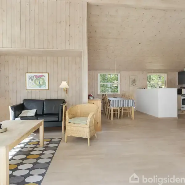 En stue med træbeklædte vægge har en sort sofa, et sofabord, og lænestol til venstre. En pejs er til højre ved det åbne køkkenområde med spisebord i baggrunden.