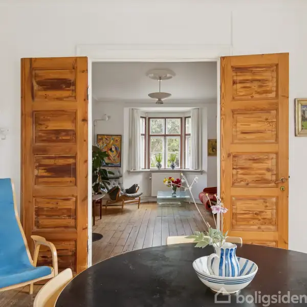 Trædør står åben og afslører en stue med trægulv, sofa og planter. Foran døren er et rundt bord med en vase og blåmalet skål. En blå stol står ved siden.