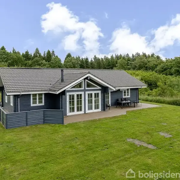 Et sort/hvidt hus står stille på en velplejet græsplæne omgivet af skov. En terrasse foran huset med havemøbler antyder et rum til afslapning.