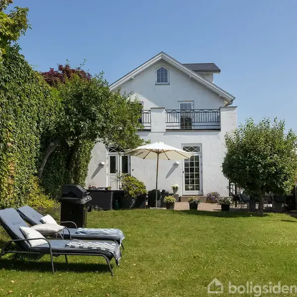 En hvid villa med balkon står bagest i en velholdt have. I haven er der solsenge, en grill og parasoller, omgivet af grønne buske og en tæt hæk.