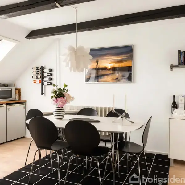 Et rundt hvidt spisebord med sorte stole står under en lamellampe. Bordet har en blomsterdekoration. I baggrunden er der et køkkenhjørne og hylder med bøger og vinflasker.