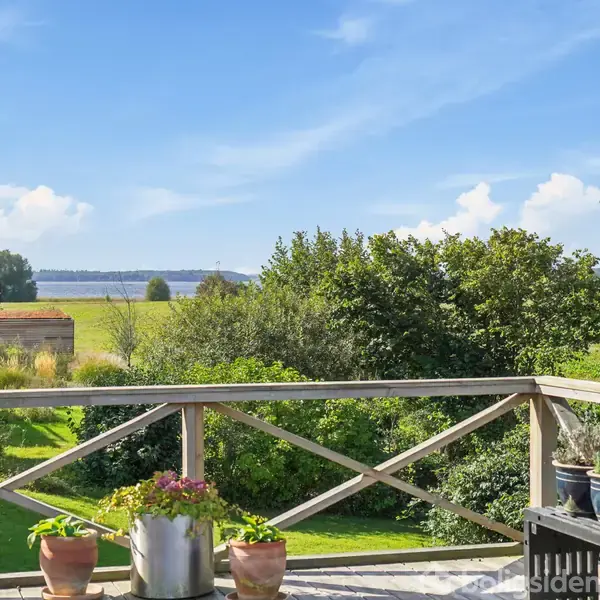 Terrasse med blomsterkrukker står stille, omgivet af grønne træer og marker, med en klar blå himmel i baggrunden. Ingen tekst er synlig.