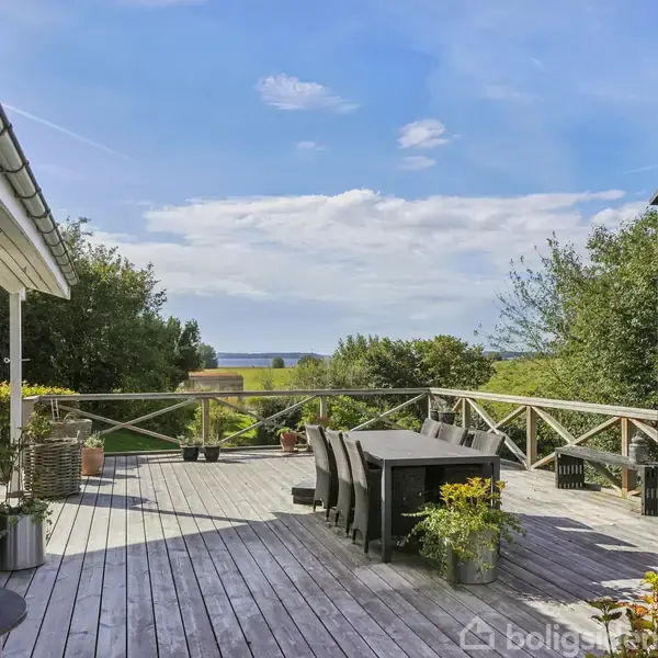 En rummelig terrasse med træmøbler og planter står ved siden af et sortmalet hus. Terrassen har udsigt over frodig natur og hav i baggrunden under en klar blå himmel.