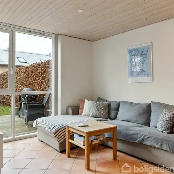 En grå sofa med farvede puder står i et lyst opholdsrum ved et sofabord. Store vinduer viser en terrasse og have udenfor. Væggen har hængt et billede.