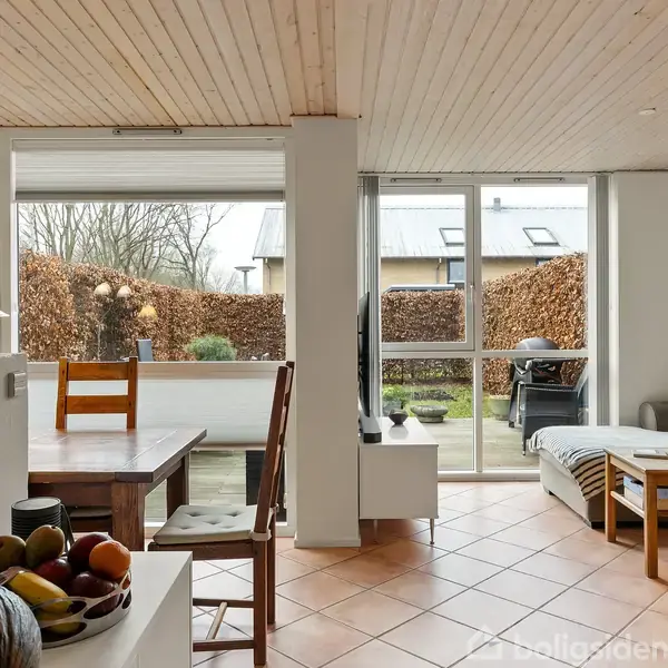 Stue med sofa, sofabord og spisebord. Rummet har lyse trælofter og terracotta-flisegulv. Store vinduer åbner ud mod en terrasse og have med træer og buske i baggrunden. Frugtskål på en hylde.