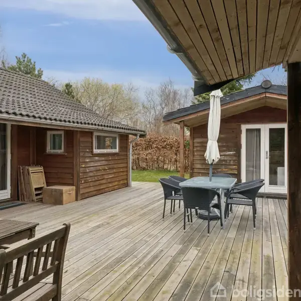 Træterrasse med møbleret bord og stole, omgivet af to træhuse med døre og vinduer. Omgivelserne inkluderer træer og buske i baggrunden.